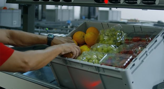 The image shows a Pick-it-Easy work station. A detailed look shows the employee placing fresh foods in crates, and preparing e-commerce orders. Oranges, grapes and tomatoes are in the crate.