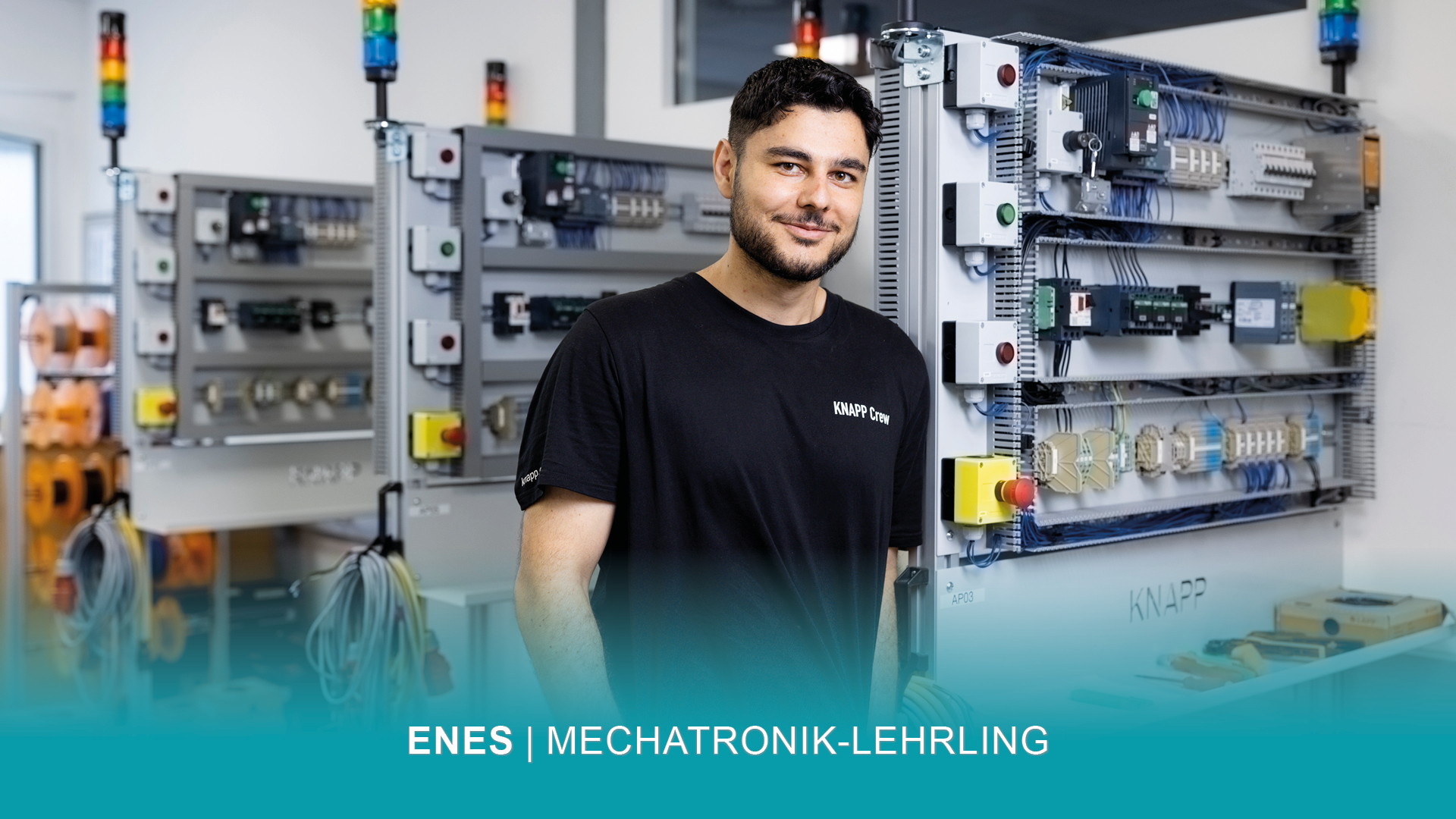 Mechatronik-Lehrling Enes steht lächelnd neben Schaltschränken in der Lehrwerkstatt