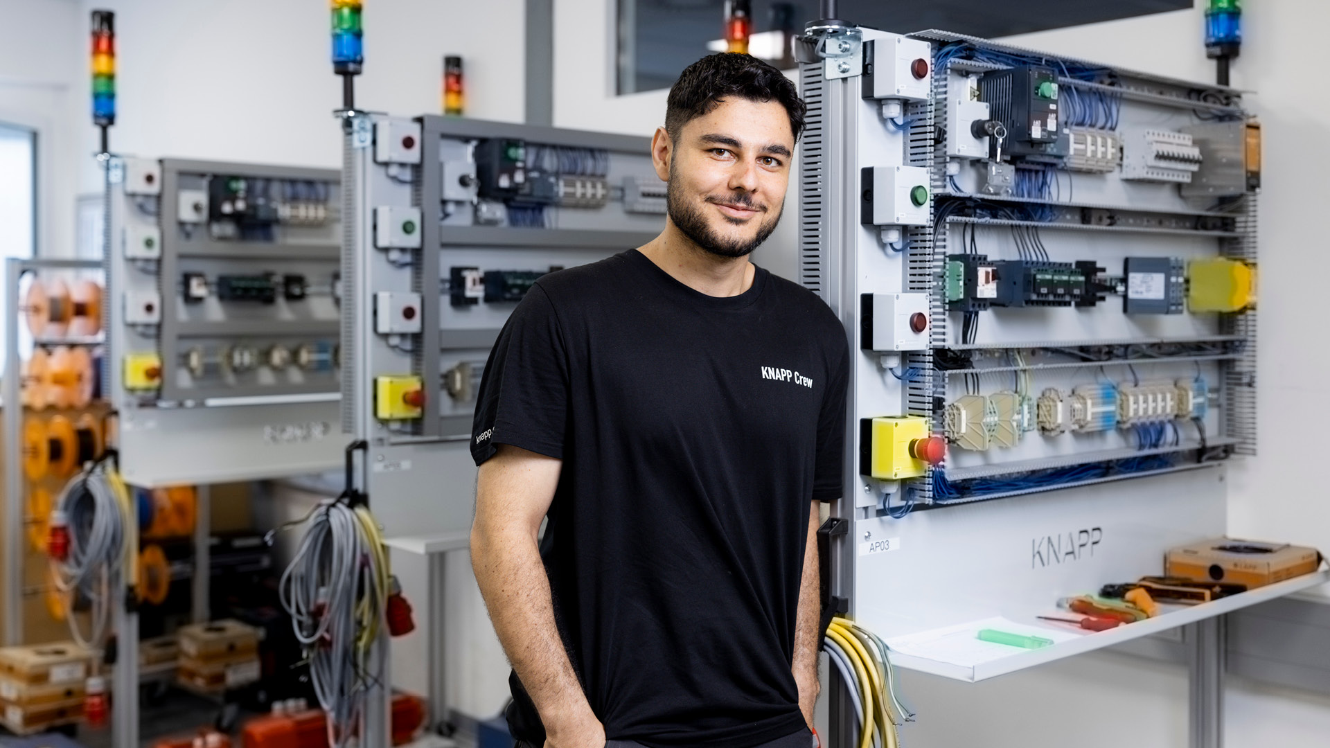 Mechatronik-Lehrling Enes steht lächelnd neben Schaltschränken in der Lehrwerkstatt