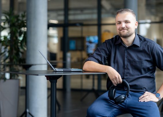 A colleague is sitting on a stool with a high table in front of him and a laptop on top. He is holding his headset loosely in his hand.