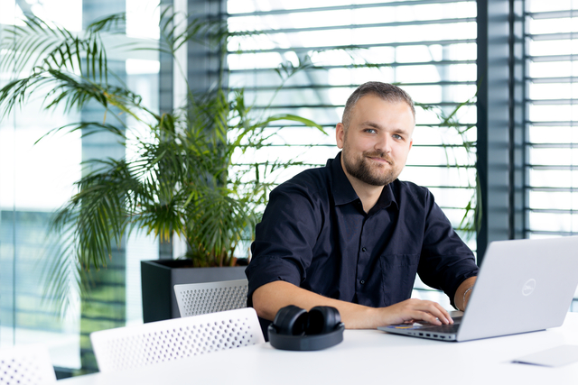 A colleague is sitting in his office. A laptop is on the table in front of him, next to it is his headset.